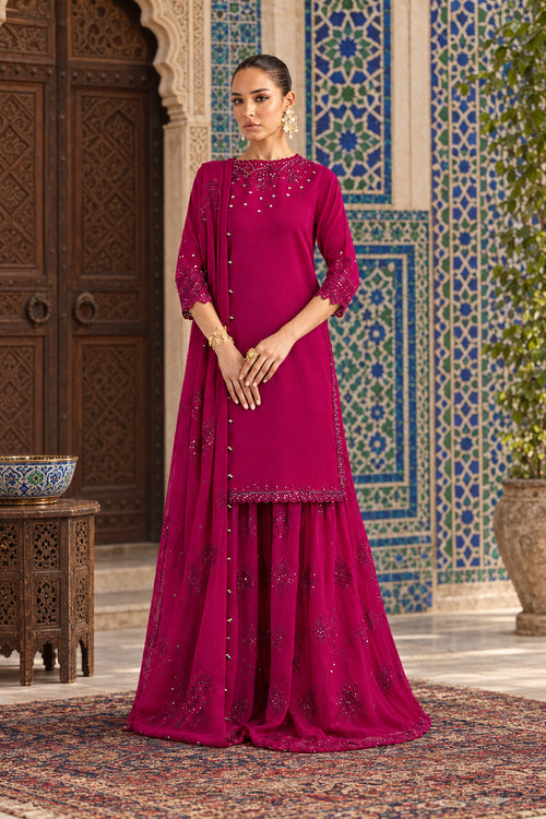 A woman models a magenta pink traditional Indian outfit consisting of a long kurta with embroidered neckline and sleeves, matching flared pants, and a draped scarf, set against a neutral gray studio background.